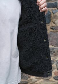 Melna vilnas jaka ar teksturētu, pūkainu materiālu, ar pogu aizdari, sānu kabatu un kontrastējošu melnu apdari.