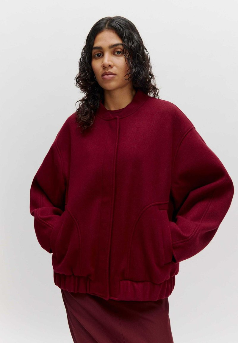 Burgunderfarbene oversized Jacke aus weichem Stoff mit rundem Halsausschnitt, Taschen vorne und geripptem Saum. Sie hat eine glatte, strukturierte Oberfläche.