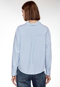 Femme aux cheveux courts bruns portant une chemise à manches longues bleu clair et un jean bleu foncé, vue de dos sur un fond blanc.