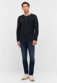 Schwarzes Langarm-Henley-Shirt mit einem geknöpften Ausschnitt, kombiniert mit dunkelblauen Jeans und beigen Sneakern, in einem lässigen Schnitt und minimalistischem Design.