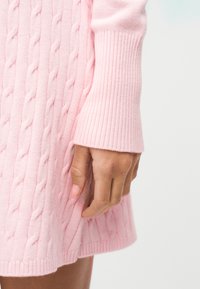 Ruka oslonjena na svijetloružičastu haljinu od pletiva s kabelskim uzorkom i rebrastim rubom na rukavu, prikazujući teksturu pletenja i detalje mekane tkanine.