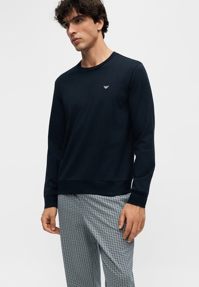 Jeune homme portant un t-shirt sombre uni à manches longues avec un petit logo et un pantalon clair à motifs, debout devant un fond uni.