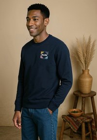 Marineblauwe sweatshirt met lange mouwen, geribbelde manchetten en een klein veelkleurig logo op de borst. Gepaard met denim jeans.