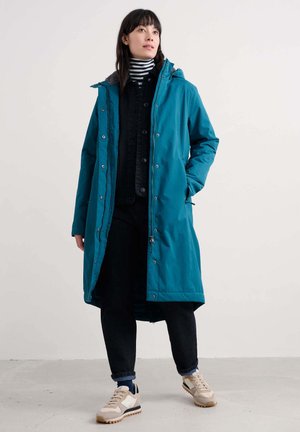 Teal lang parka med hætte, der har lynlås og knaplukning. Sammen med en sort jakke, denimbukser og beige sneakers. Stribet skjorte under.
