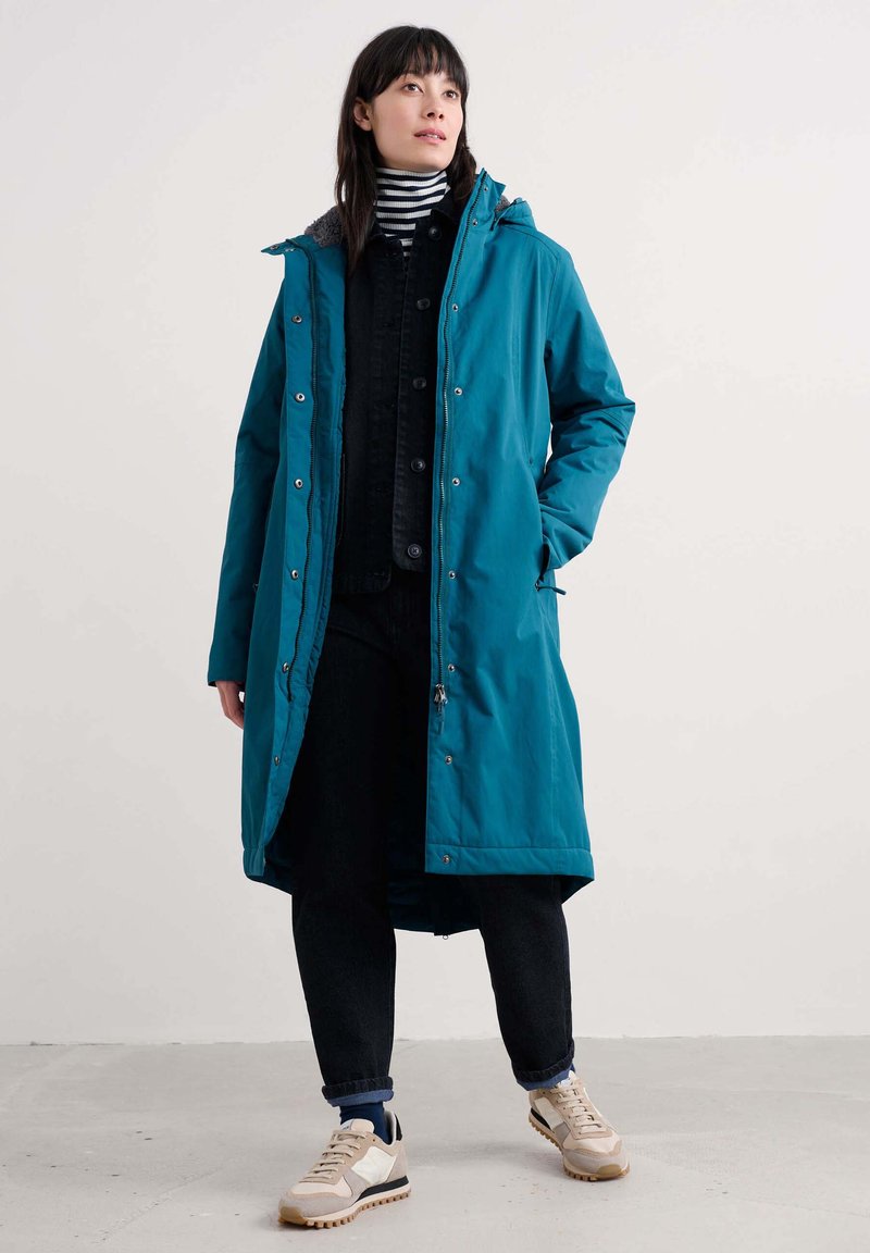 Parka lunga teal con cappuccio, dotata di zip e bottoni automatici. Abbinata a una giacca nera, pantaloni di jeans e sneakers beige. Camicia a righe sotto.