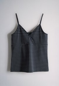 Black sleeveless top with white polka dots, V-neck, and thin adjustable shoulder straps hanging against a light gray background.