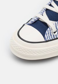 Blå kanvas sneakers med hvit gummitå, teksturert såle, svart gummilist, og kontrastfargede stripete snørebånd.