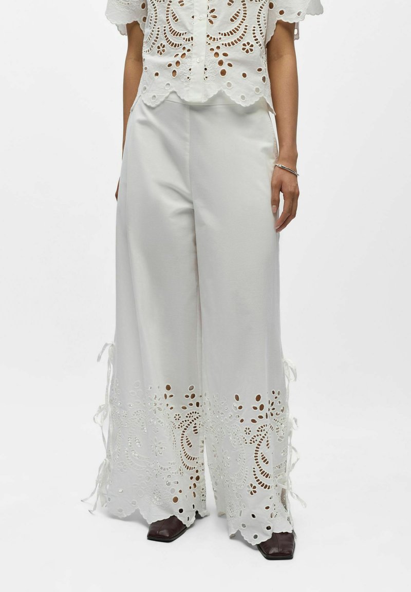 Femme portant un pantalon blanc à jambes larges avec des découpes florales ajourées et des liens sur les côtés, assorti à des chaussures marron à bout carré et un haut en dentelle blanche.