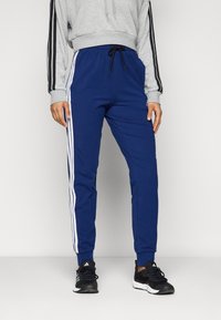 Blå joggarbyxor med vita sidostränder, elastisk midja och ribbade muddar. Kombineras med en grå kort sweatshirt och svarta träningsskor.
