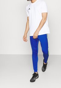 Man med vit Adidas T-shirt, blå träningsbyxor med neonlysande rand och svarta fotbollsskor som står mot en enfärgad bakgrund.