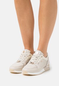 Gambe con scarpe sportive beige stringate, con pannelli in mesh e pelle, dettagli dorati e suola bianca, su sfondo bianco.