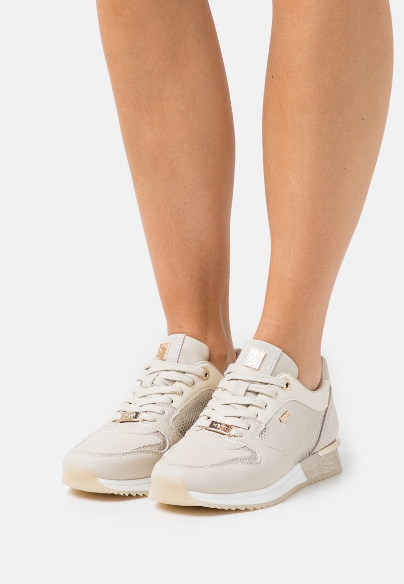 Gambe con scarpe sportive beige stringate, con pannelli in mesh e pelle, dettagli dorati e suola bianca, su sfondo bianco.