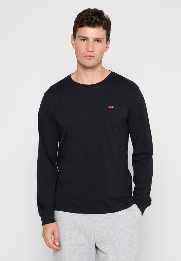 Camisa negra de manga larga hecha de algodón, con un cuello redondo y un pequeño logo rojo de Levi's en el pecho. Lleva puesta con pantalones de chándal grises.
