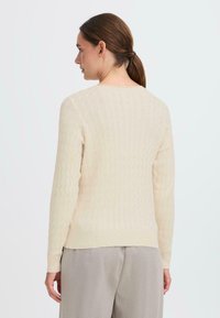 Gebreide trui in lichtcrème met een gestructureerd ruitpatroon, lange mouwen en een ronde halslijn. Gecombineerd met lichtgrijze trousers.