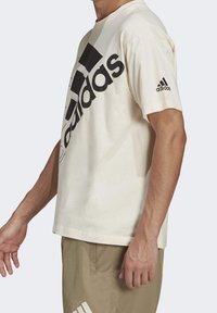 Camiseta de algodón crema con un gran logo negro de "adidas" y tres franjas negras en la parte frontal. Mangas cortas, corte relajado, diseño minimalista.