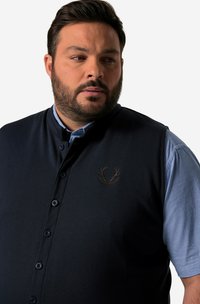 Mörkblå ärmlös väst med knäppning, med rund krage och en brun broderad logotyp på bröstet. Under är en blå rutig skjorta.