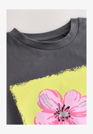 Siva pamučna majica s okruglim izrezom, s velikim ružičastim cvijetom obrubljenim crnom na svijetlo žutoj pozadini.