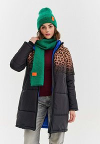 Žena v čiernom prešívanom kabáte s leopardím vzorom, zelenej pletenej čiapke, zodpovedajúcom zelenom šále, vínovej blúzke a svetlo šedých džínsach.