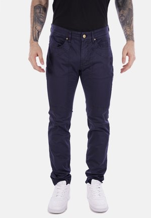 Pantaloni in misto cotone blu navy con taglio slim, dotati di cinque tasche, zip e cuciture resistenti. Indossati con sneaker bianche.