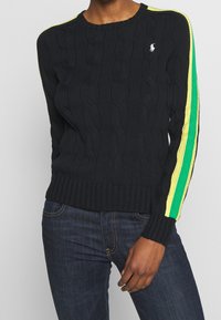 Person som bär en svart kabelstickad tröja med gula och gröna ränder på ärmarna och en vit logotyp på bröstet, tillsammans med mörkblå jeans.