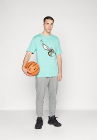 Tirkīza krāsas T-krekls ar bitenes grafiku un basketbola bumbu, pelēkas sporta bikses un melni apavi, stāv uz baltā fona.