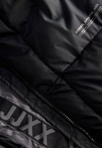 Melna quiltota jaka ar redzamu rāvējslēdzēju, kas rotāta ar balto tekstu "DESIGN APPAREL JJXX" un zīmola iekšējo oderi, kurā atkārtojas "JJXX".
