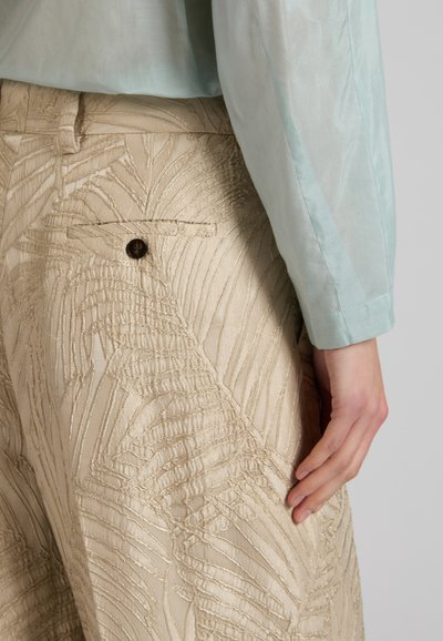 Pantaloni beige con motivo a foglia testurizzato e tasca posteriore con bottone, abbinati a una camicia a maniche lunghe azzurro chiaro e mano appoggiata di lato.
