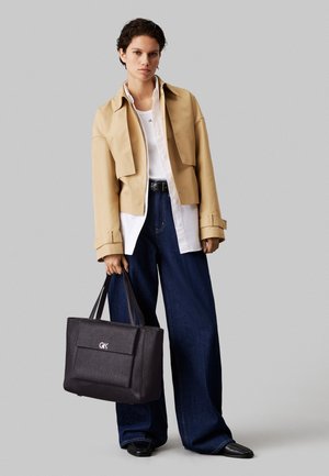 Zwarte tote bag van gestructureerd materiaal met een voorkantzak, vastgehouden in een hand; persoon draagt een beige cropped jack over gelaagde tops en wijde jeans.