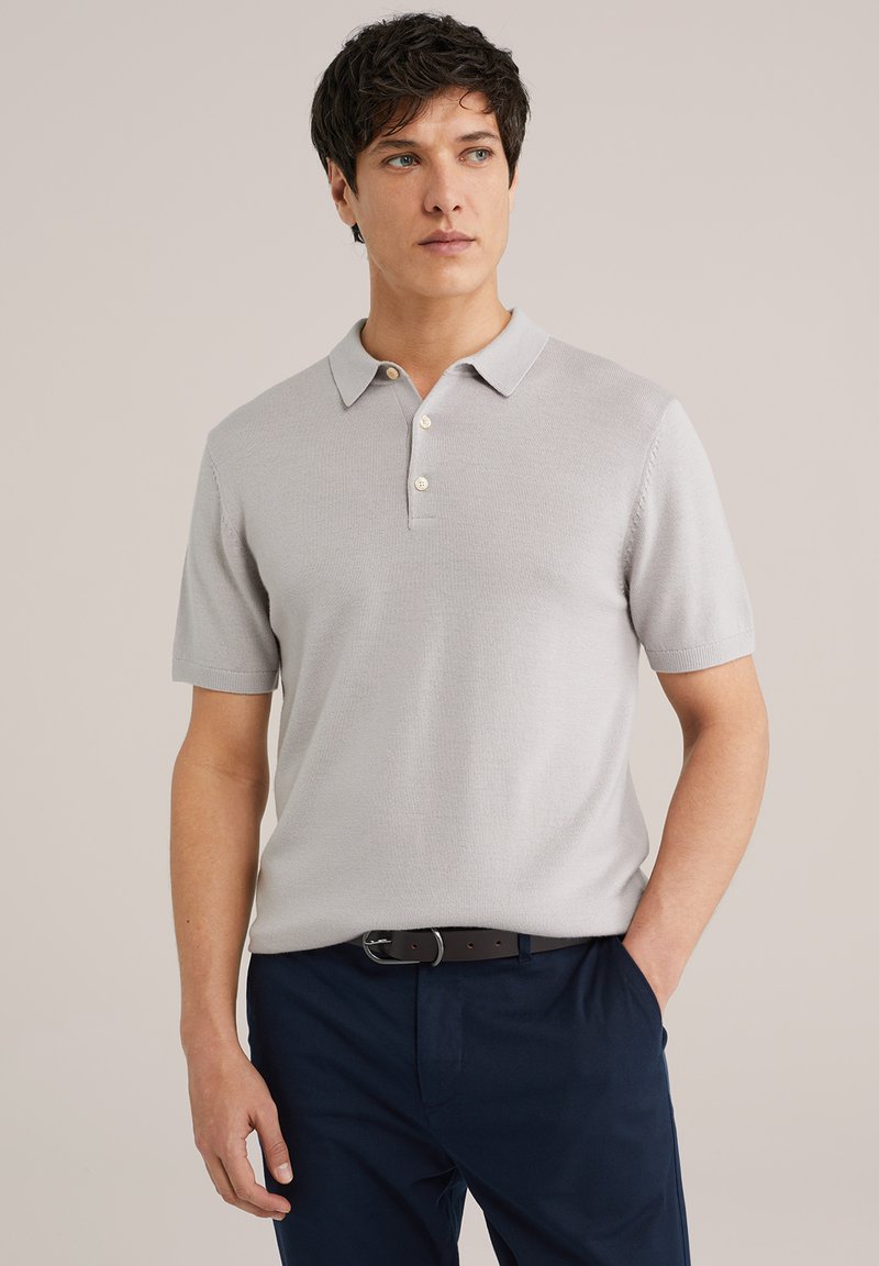 Gaiši pelēks īsās piedurknes polo krekls ar strukturētu adījumu, ar klasisku apkakli un trim pogām, savienots ar tumši ziliem biksēm.