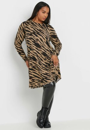 Langärmliges braunes Kleid mit schwarzen Tigermustern; textil strukturiertes Gewebe, leicht ausgestellter A-Linien-Schnitt, mit einer wellenförmigen Saumabschlüssen. Getragen mit schwarzen Leggings und Stiefeln.