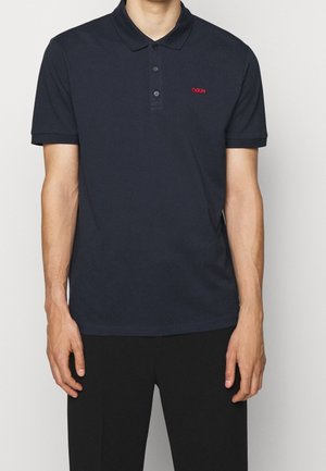 Man draagt een donkerblauw poloshirt met korte mouwen en een rood "HUGO"-logo op de linkerborst, gecombineerd met een zwarte broek.