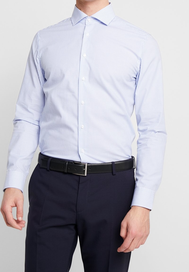 Homme portant une chemise à rayures bleu clair et blanc boutonnée, rentrée dans un pantalon bleu marine foncé avec une ceinture noire.