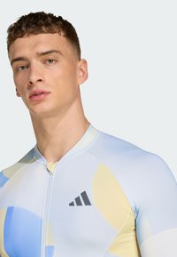 Herren-Sportanzug in Hellblau und Pastellgelb, mit einem Reißverschluss, kurzen Ärmeln und schwarzen Logo-Akzenten. Glatter, dehnbarer Stoff.