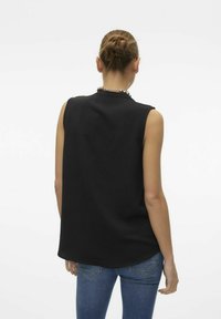 Haut sans manches noir avec une surface texturée, décolleté rond et coupe ample, associé à un jean bleu. Cou décoré d'un collier en perles.