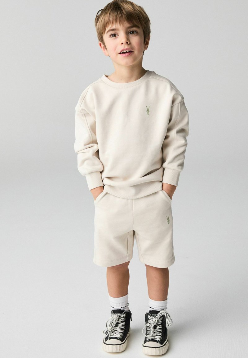 Beige bomuldssweater med grøn broderi, parret med matchende shorts. Sorte høj-top sneakers med hvide snørebånd og ribbede sokker.