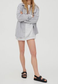 Grå skjorte med knap, lys grå sweatshirt, grå shorts og sorte sandaler med knude detaljer. Enkel, afslappet lagdelt outfit med bløde teksturer.
