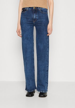 Persoon in hoge taille, rechte lichtblauwe spijkerbroeken en zwarte hoge hakken, staand tegen een effen lichte achtergrond.