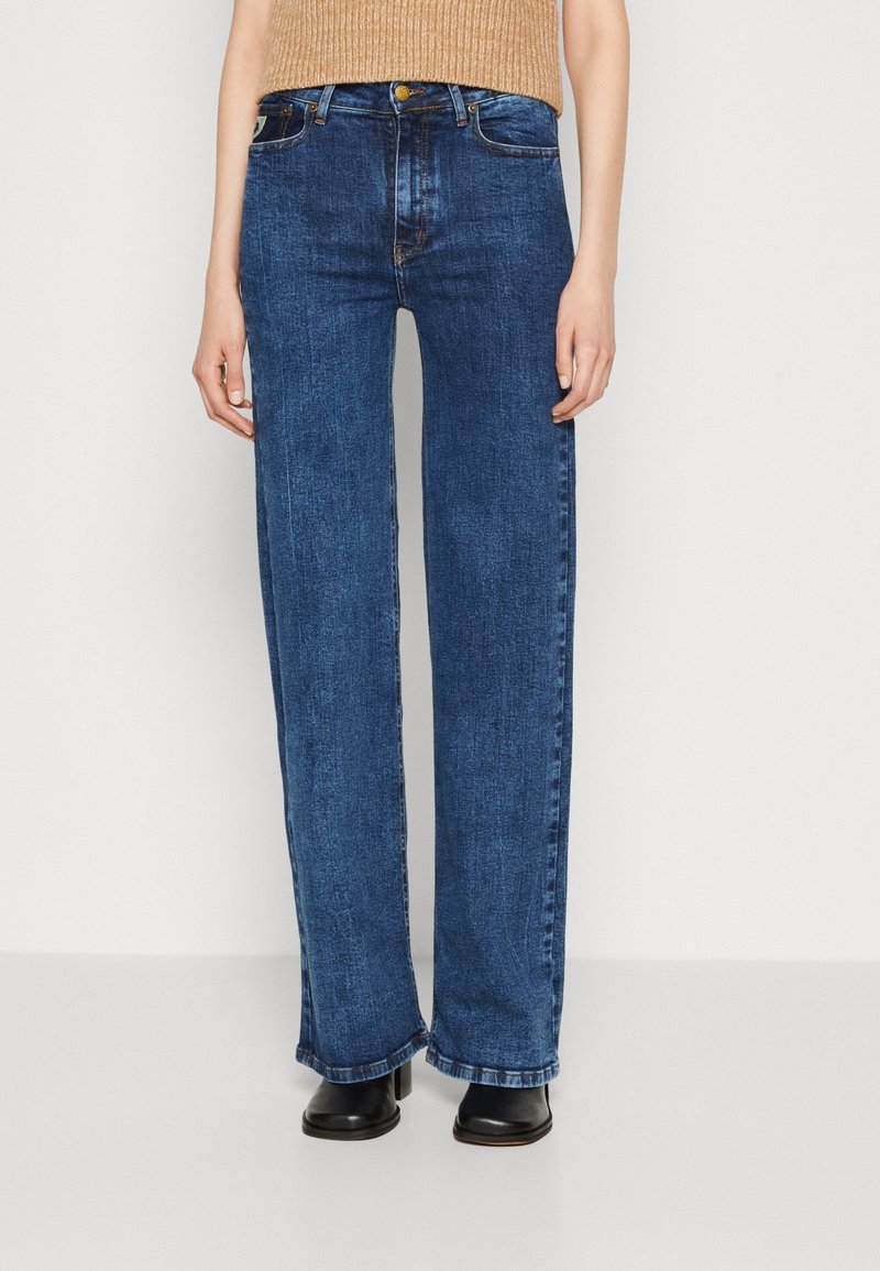 Henkilö, joka on pukeutunut korkeavyötäröisiin, suoriin sinisiin farkkuihin ja mustiin korkokenkiin, seisoo yksinkertaista vaaleata taustaa vasten.