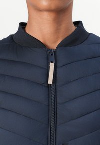 Marineblå polstret jakke med høy krage, glidelåslukking og chevron-quilting. Har en kontrastfarget glidelåspull og teksturert krage.