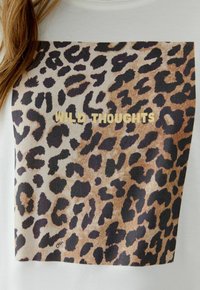T-shirt blanc avec un grand motif léopard et le texte doré "WILD THOUGHTS" en police gras, au design texturé et avec des accents sombres.