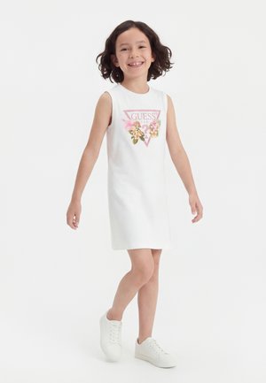 Fille souriante marchant, portant une robe blanche sans manches avec un logo floral Guess et des baskets blanches sur un fond blanc uni.