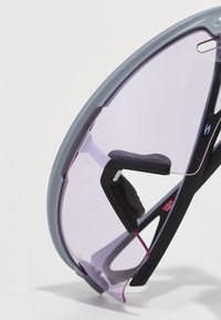 Sonnenbrille mit einem grauen Kunststoffrahmen, lila getönten Gläsern und schwarzen Gummiakzenten. Hat ein sportliches Design mit offenen Bügeln und verstellbaren Nasenpolstern.