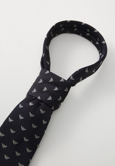 Laivastonsininen solmio teksturoidusta kankaasta, jossa on toistuva hopeinen logo-kuvio. Sisältää tavallisen solmun ja puhtaan, suppenevan pään.