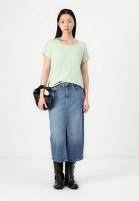 Hellgrünes Kurzarm-T-Shirt, kombiniert mit einem blauen Denim-Rock mit Schlitz vorne. Hält eine kleine schwarze Handtasche. Schwarze knielange Stiefel.