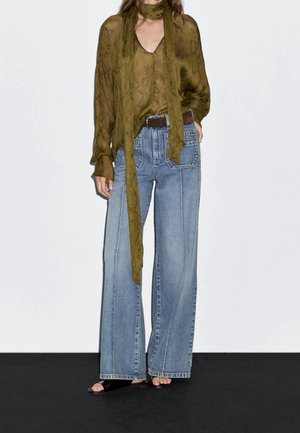 Personne portant un chemisier transparent vert olive avec une longue écharpe, un jean large bleu clair avec une ceinture marron et des sandales noires ouvertes aux orteils.