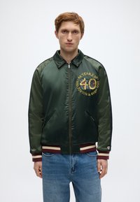 Πράσινο σατέν μπουφάν bomber με κεντημένο "40" και λογότυπο. Διαθέτει ριμπ μανσέτα, γιακά και τελείωμα σε βουργουνδί και εκρού χρώμα, φερμουάρ και πλαϊνές τσέπες.