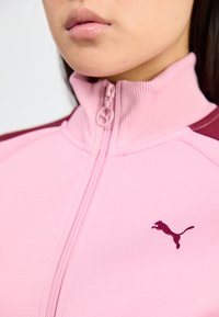 Nærbillede af en person iført en lyserød zip-up jakke med en maroon streg og et lille mørkt logo af en springende kat på brystet.