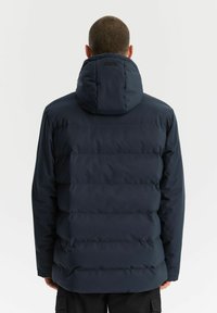 Mężczyzna z krótkimi włosami, noszący ciemnoniebieską ocieplaną zimową kurtkę z kapturem, pokazany od tyłu na jednolitym tle.