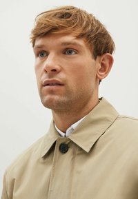 Beige trenchcoat med en knaplukket krave. Stoffet ser glat ud med en let glans. Modellen har kort, lysebrunt hår og blå øjne.
