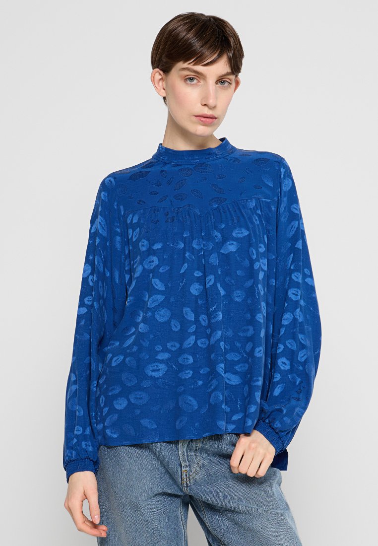 Frau mit kurzen braunen Haaren, die eine locker sitzende blaue Bluse mit langen Ärmeln und einem subtilen Lippenmuster trägt, sowie hellblauen Jeans.